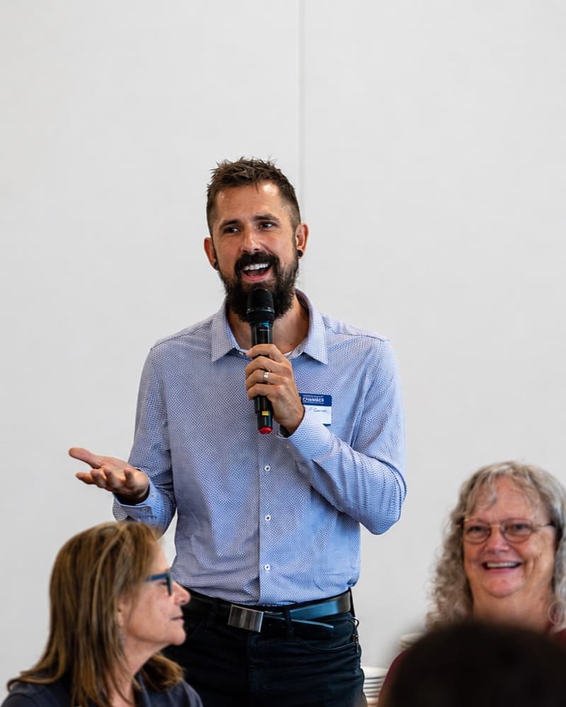 Alexander Gartley speaking into a microphone with an open-hand gesture at the Greater EMC Chamber Great 88 breakfast